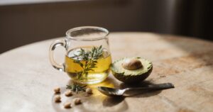 Avocado-Öl in Glasflasche mit halbierter Avocado und Kräutern auf Holztisch in Küche