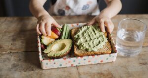 Bunte Pausenbox mit Avocado-Snacks, Gemüse-Sticks und Obst auf Holztisch, Kinderhand greift zu