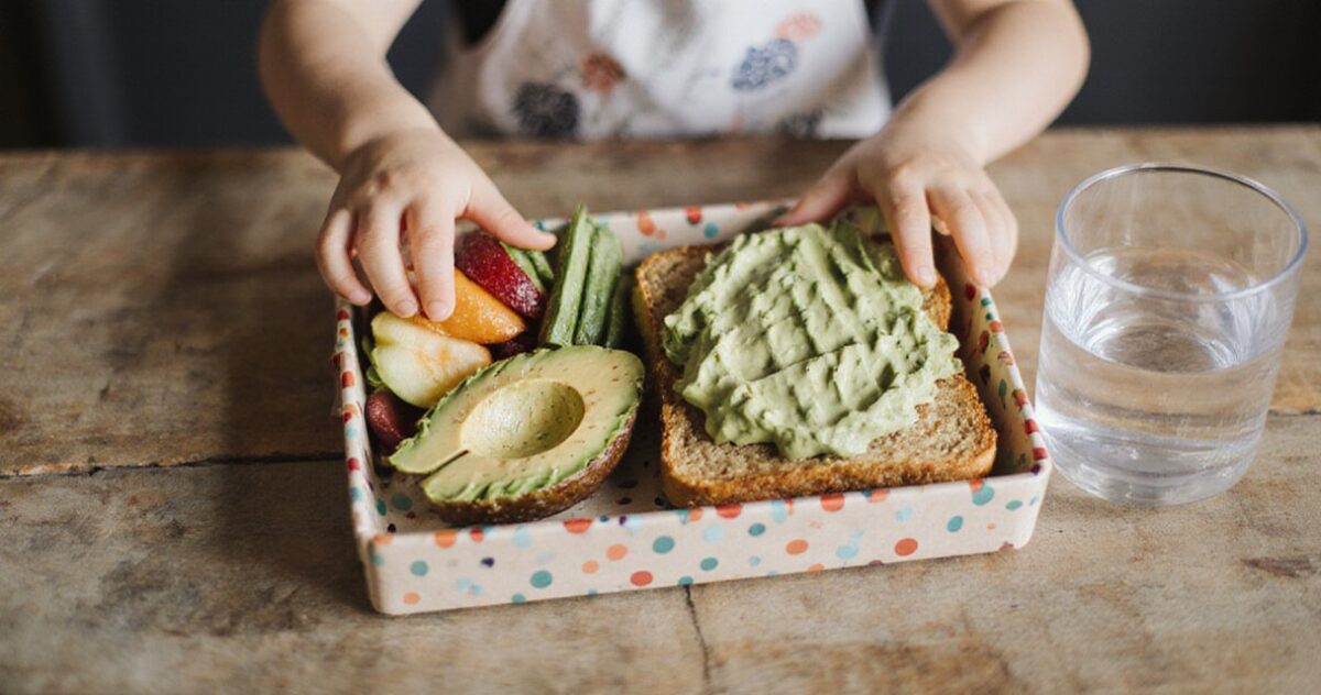 Bunte Pausenbox mit Avocado-Snacks, Gemüse-Sticks und Obst auf Holztisch, Kinderhand greift zu